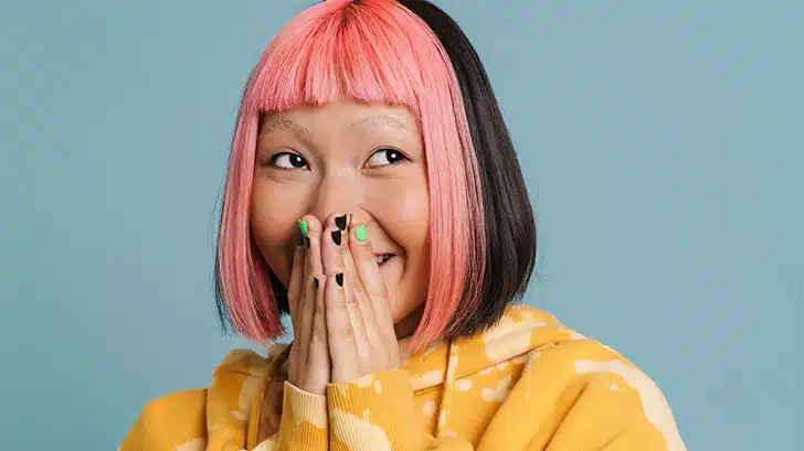Woman with pink hair, cut in a bob style smiles with hands over mouth.