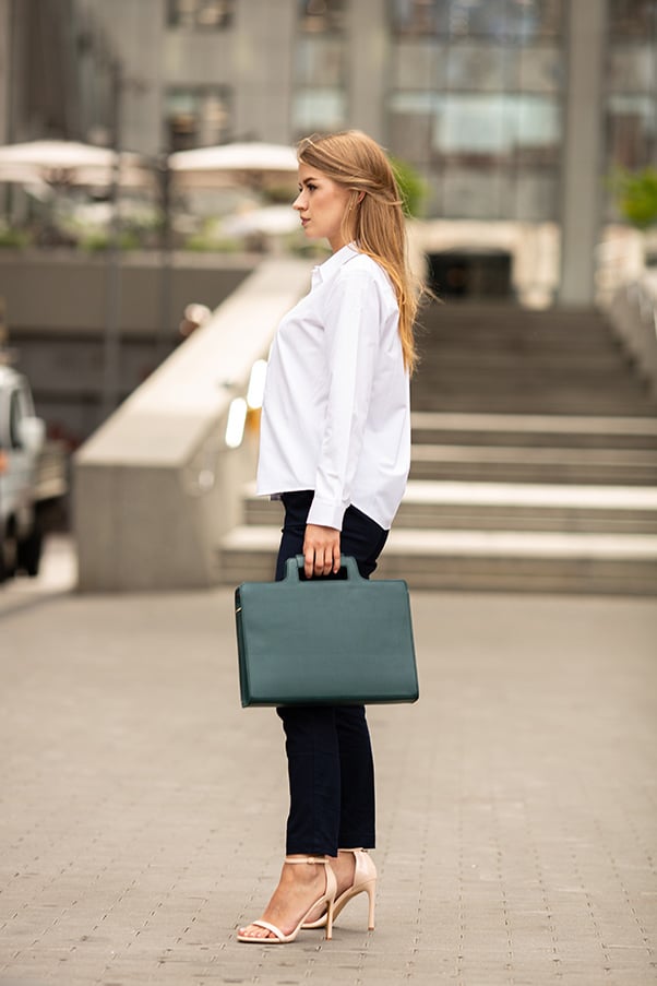 Woman wearing white button down top and black pants, carrying green leather briefcase.