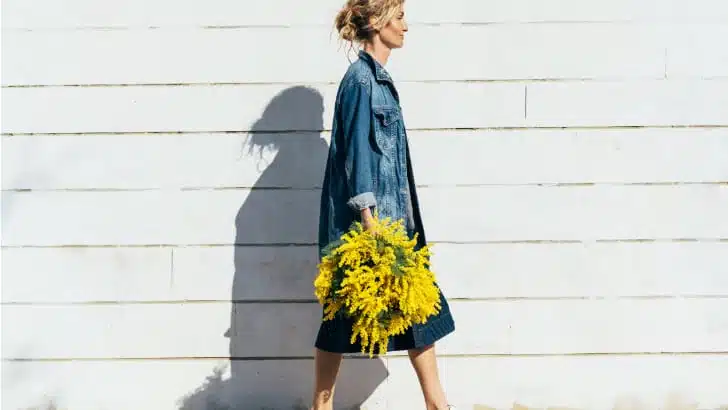 Woman wearing fall denim pieces walks in front of wall holding flowers.